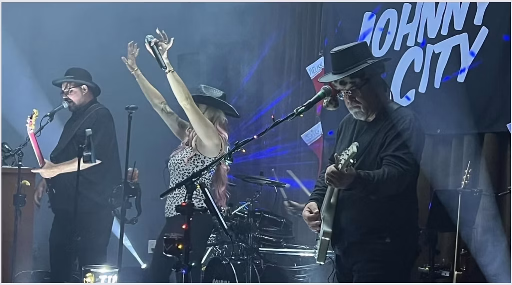 Three musicians perform on stage with instruments and hats; “JOHNNY CITY” is visible on the background wall.