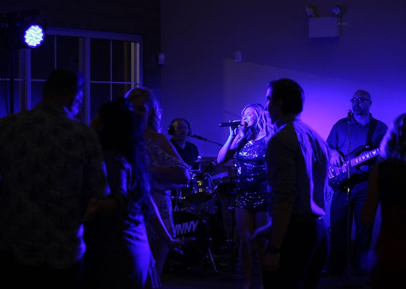 A female vocalist performs on stage with a band while people dance in a dimly lit room with blue lighting.