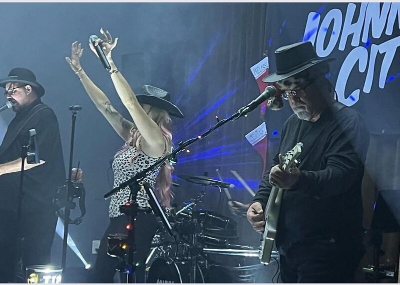 A band performs under blue lights; three members in hats play instruments with a “JOHNNY CITY” sign behind them.