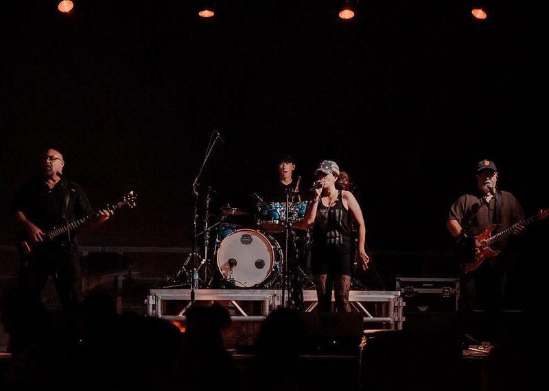Four musicians perform on stage under orange lights, featuring a singer, drummer, and two guitarists.