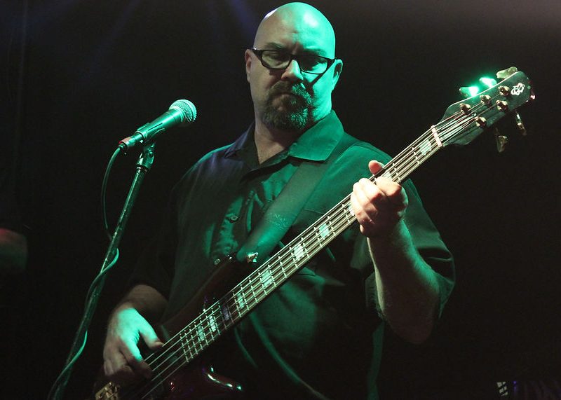 Man playing an electric bass guitar on stage near a microphone, wearing glasses and a dark shirt under stage lighting.