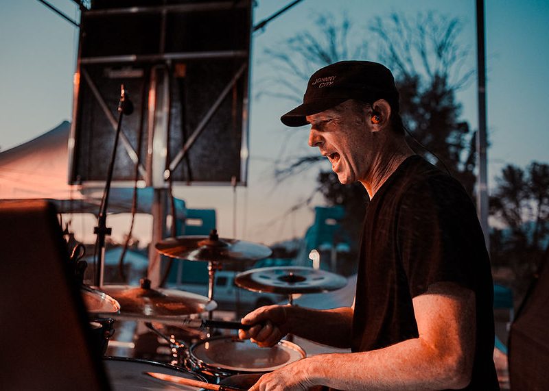 Man in black shirt plays drums outdoors at sunset, with trees and stage equipment visible in the background.