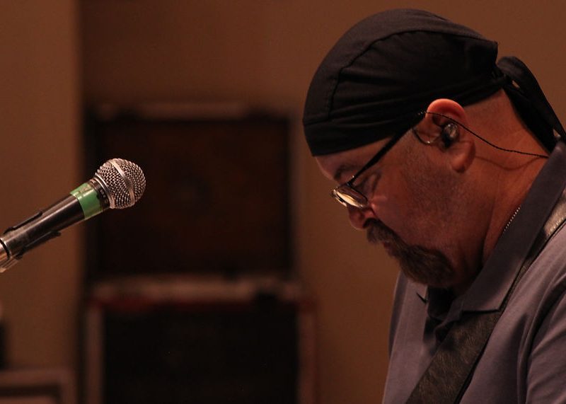 Man wearing a headscarf and glasses plays guitar near a microphone, looking down with in-ear monitors.