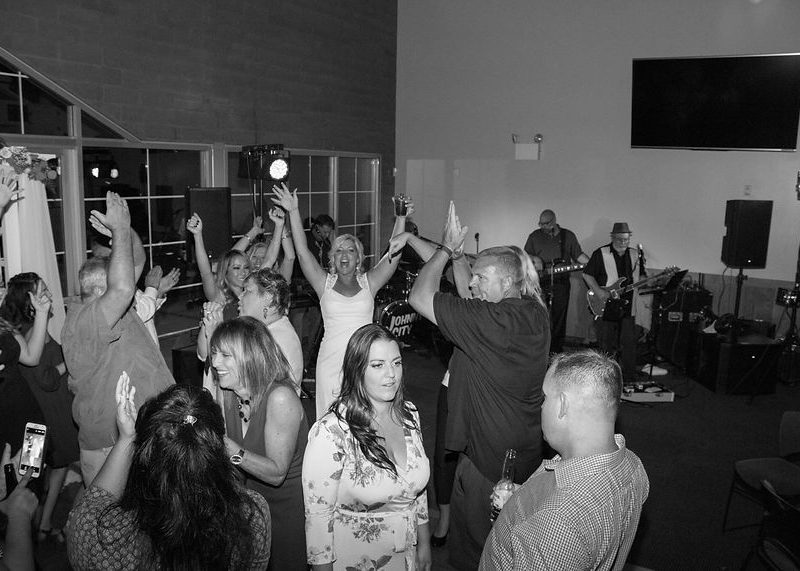 People dance and celebrate in a lively indoor party while a band performs on stage in the background.