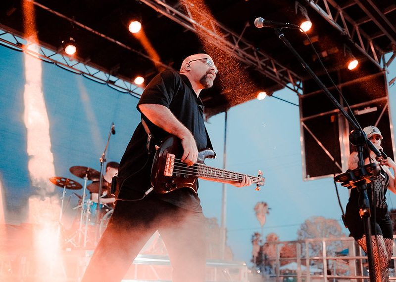 Bassist in black shirt performing on outdoor stage with band, orange stage lights, and evening sky in background.