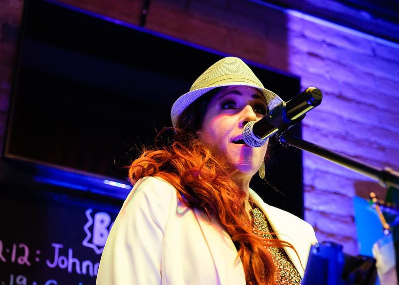 Person in a white hat and jacket singing or speaking into a microphone on a stage with blue and purple lighting.