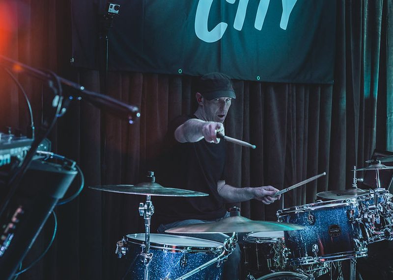 A drummer wearing a cap plays a drum set on stage, pointing a drumstick forward while performing.
