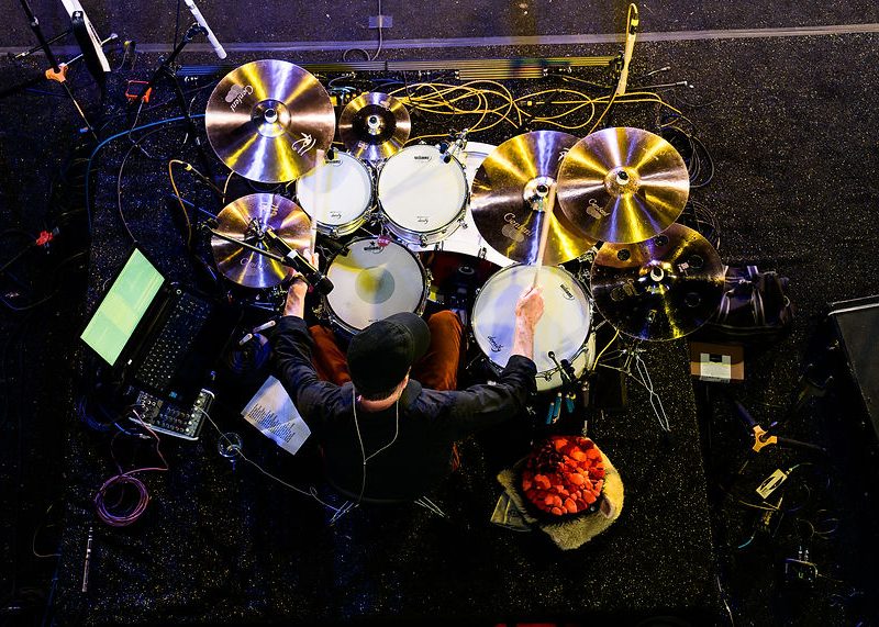 A drummer plays a drum kit surrounded by cymbals, cables, and electronic equipment, viewed from above.